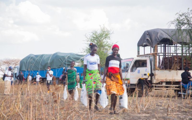 The image shows two individuals walking through a dry, harvested field while carrying large bags of supplies. Several trucks and aid vehicles are parked nearby, with other people gathered around the distribution site. The scene reflects a humanitarian assistance operation, where relief items are being delivered and collected by local community members in a rural setting. The cloudy sky and open landscape emphasize the outdoor nature of the event and the logistical effort involved in providing support to the area.